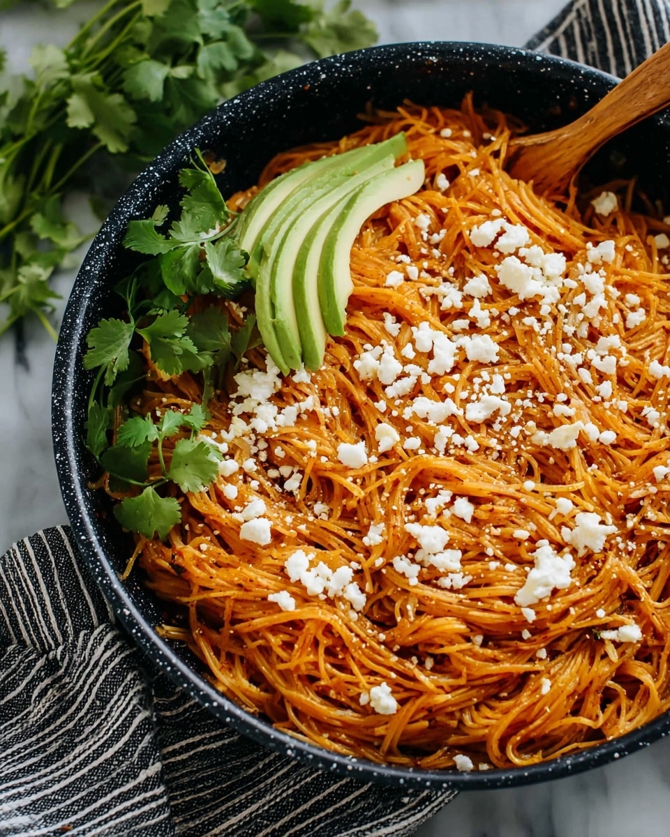 A close-up shot of a black speckled pan filled with thin, orange-red noodles layered evenly across, topped with crumbled white cheese scattered over the noodles. On one side, there are two slices of light green avocado fanned out neatly on top of the noodles, while fresh green cilantro leaves rest next to the avocado on the edge of the pan. The pan sits on a white and black striped cloth, placed on a surface with a white marbled texture. A wooden utensil handle is visible resting on the noodles at the back, photo taken with an iphone --ar 4:5 --v 7