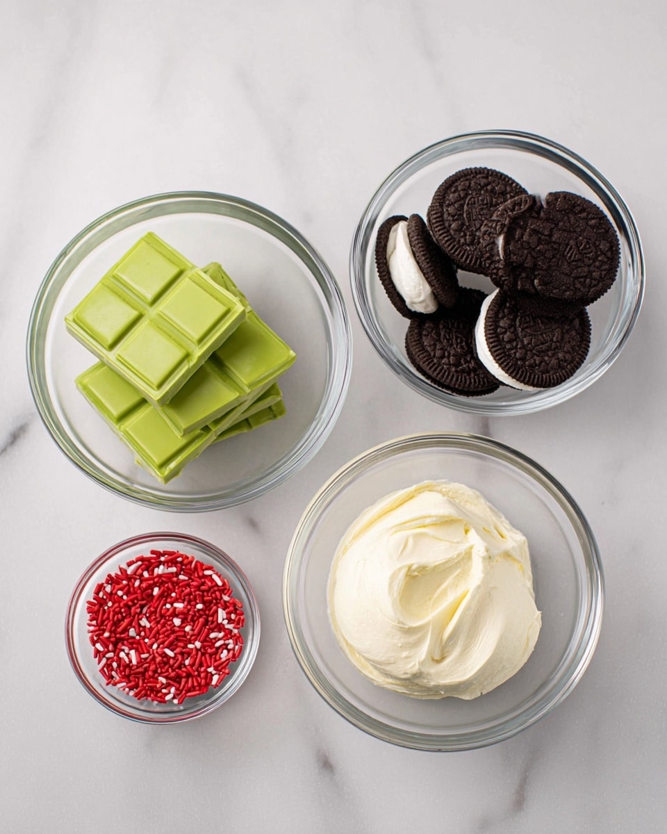 Four clear glass bowls are placed on a white marbled surface. The top left bowl holds three thick, bright green bars of chocolate stacked neatly. The top right bowl is filled with round, dark brown sandwich cookies with cream filling, arranged loosely. The bottom left bowl contains a creamy, soft white spread with a swirled texture. In front of these bowls, there is a small clear bowl filled with small red heart-shaped sprinkles. photo taken with an iphone --ar 4:5 --v 7