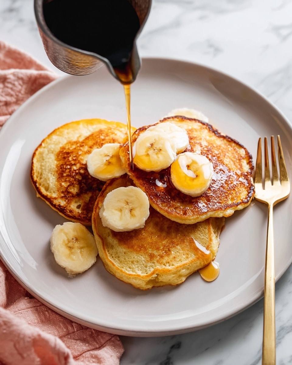 Three golden brown pancakes are stacked slightly overlapping on a white plate with a soft edge design. There are five round banana slices placed on top and beside the pancakes. A dark metal pitcher is pouring amber syrup over the middle pancake, creating a shiny pool. A gold fork rests on the right edge of the plate. The background is a white marbled surface with a soft pink cloth on the bottom left corner. photo taken with an iphone --ar 4:5 --v 7