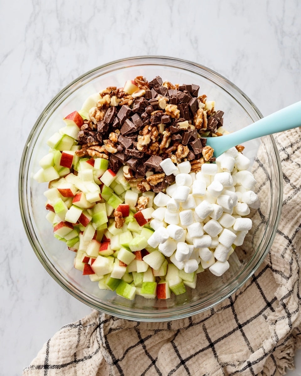 A clear glass bowl with three layers of ingredients is placed on a white marbled surface beside a beige and black checkered cloth. The bottom layer is made of small, chopped pieces of green and red apples. On top of this is a layer of chocolate chunks with a mix of dark brown and light tan colors showing the nut pieces inside. The top layer is a pile of small white mini marshmallows. A light blue spoon is resting on the right edge of the bowl, partially submerged in the marshmallows. Photo taken with an iphone --ar 4:5 --v 7