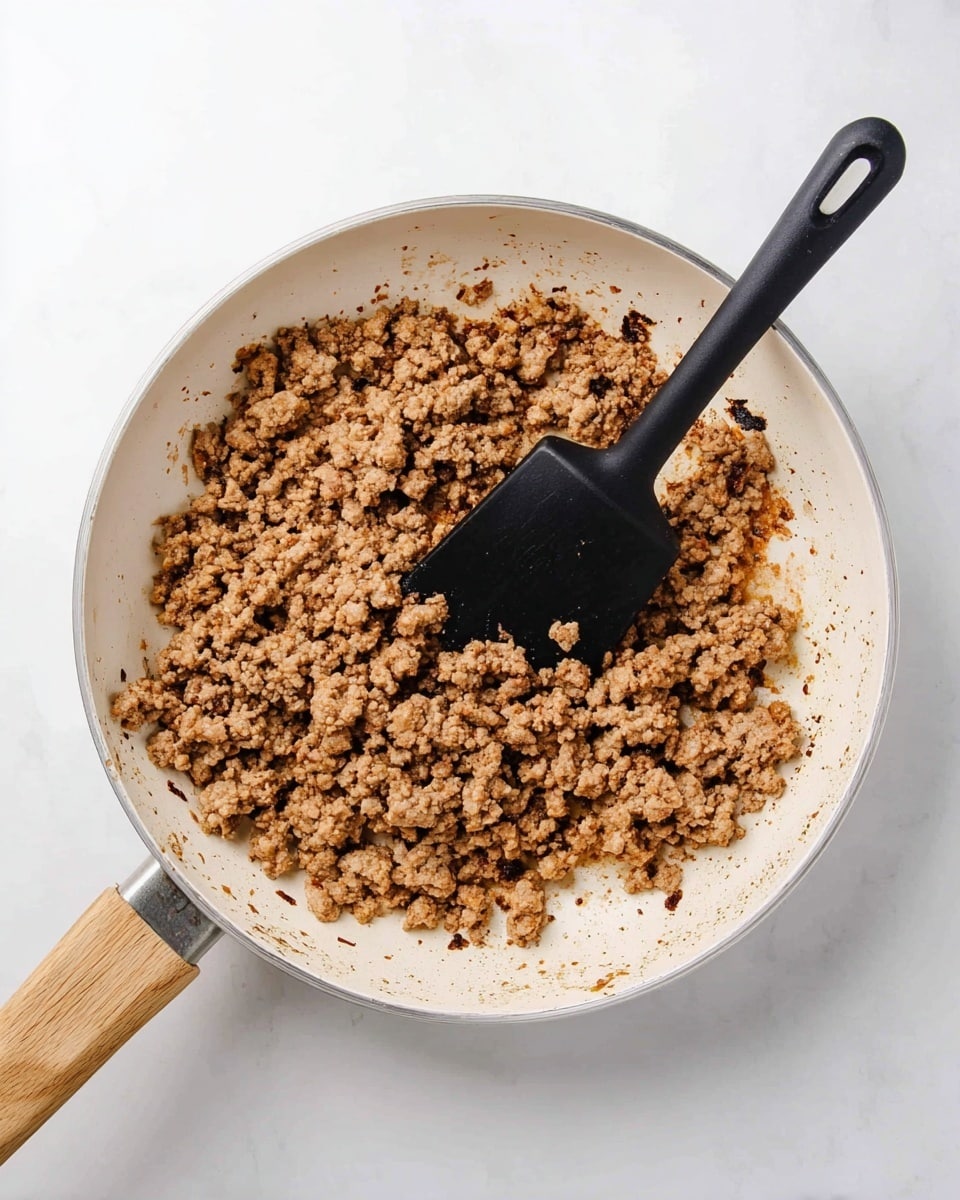 A top view of a white frying pan with a light wooden handle, filled with cooked ground meat that is light brown with some darker brown bits, showing a crumbly texture, with browned edges sticking slightly to the pan. A black spatula with a matte handle rests in the pan, partly over the meat. The pan sits on a white marbled surface that is smooth and clean. photo taken with an iphone --ar 4:5 --v 7
