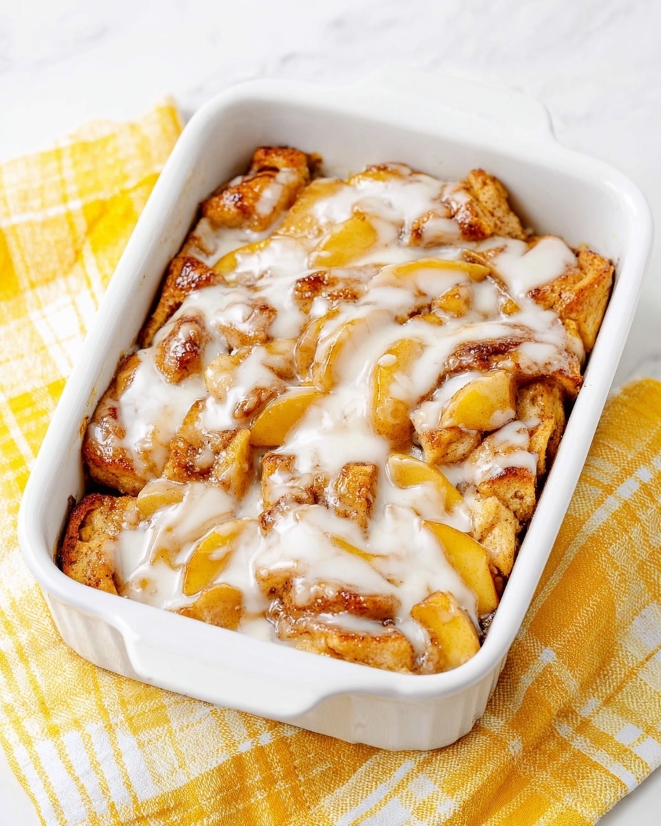 The image shows a white rectangular baking dish filled with a warm dessert that has several layers. The base layer consists of soft, golden-brown pastry pieces with some darker spots from baking. Scattered over the base are tender, cooked apple slices in light yellow and caramel colors mixed with a glossy syrup. The top layer is a smooth, white icing drizzle that spreads unevenly over the dessert, adding a creamy texture contrast. The dish sits on a yellow and white checked cloth placed on a white marbled surface. photo taken with an iphone --ar 4:5 --v 7