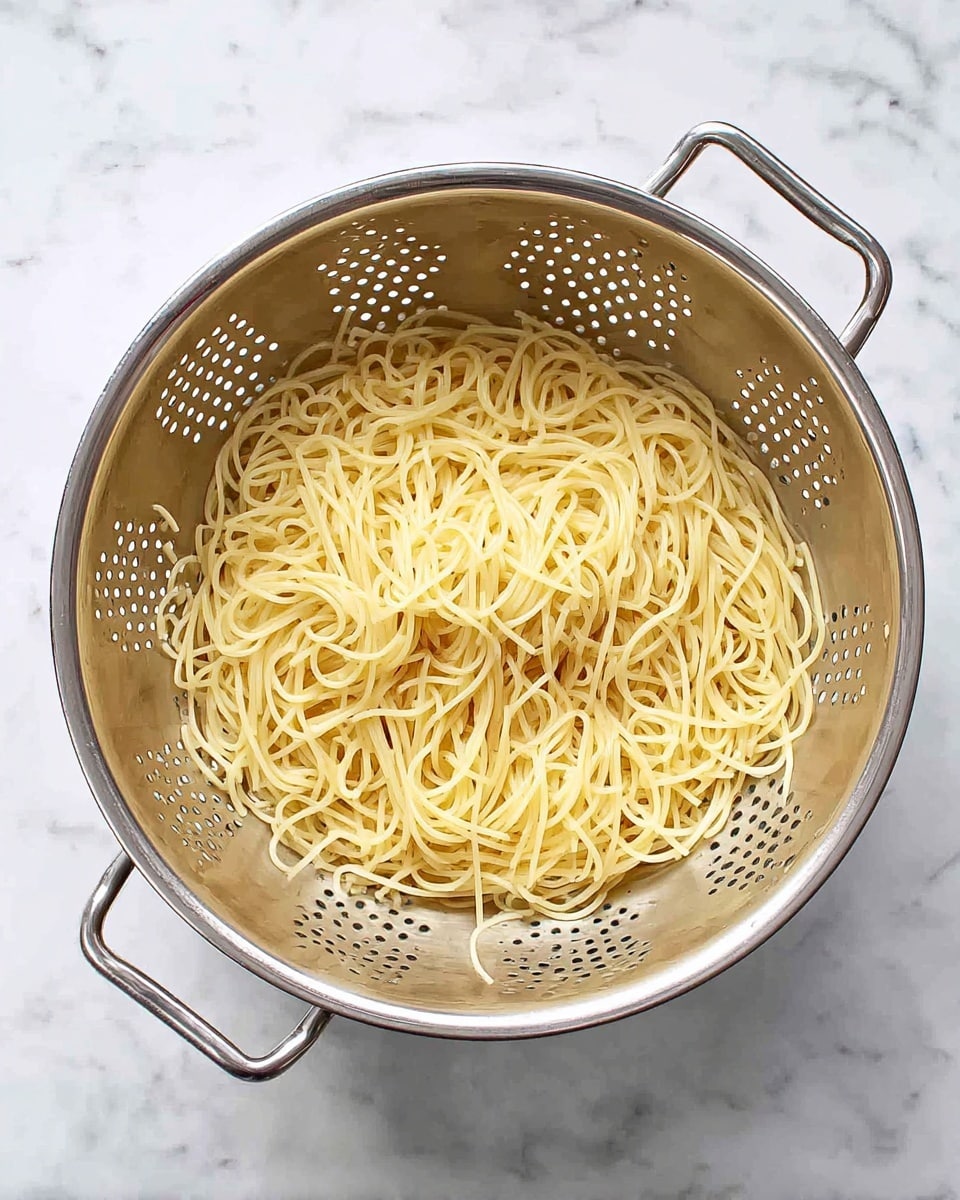 A large silver metal colander filled with cooked thin spaghetti pasta. The pasta is light yellow and slightly shiny, resting evenly at the bottom inside the colander. The colander has many small round holes and two handles on each side. It is placed on a white marbled surface. photo taken with an iphone --ar 4:5 --v 7