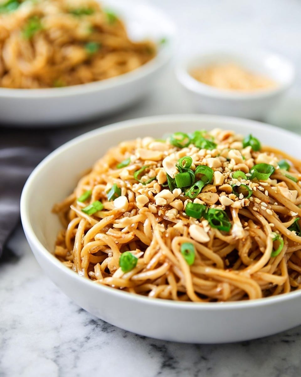 A white bowl filled with light brown noodles sits in the foreground, topped with chopped green onions, crushed peanuts, and sprinkled sesame seeds. The noodles have a slight shine and a smooth texture, with the green onions scattered evenly on top, adding bright green pops of color. The crushed peanuts provide a rough texture and pale tan color contrast. In the blurred background, there is another white bowl with more noodles and a small white bowl filled with sesame seeds placed on a white marbled surface. photo taken with an iphone --ar 4:5 --v 7