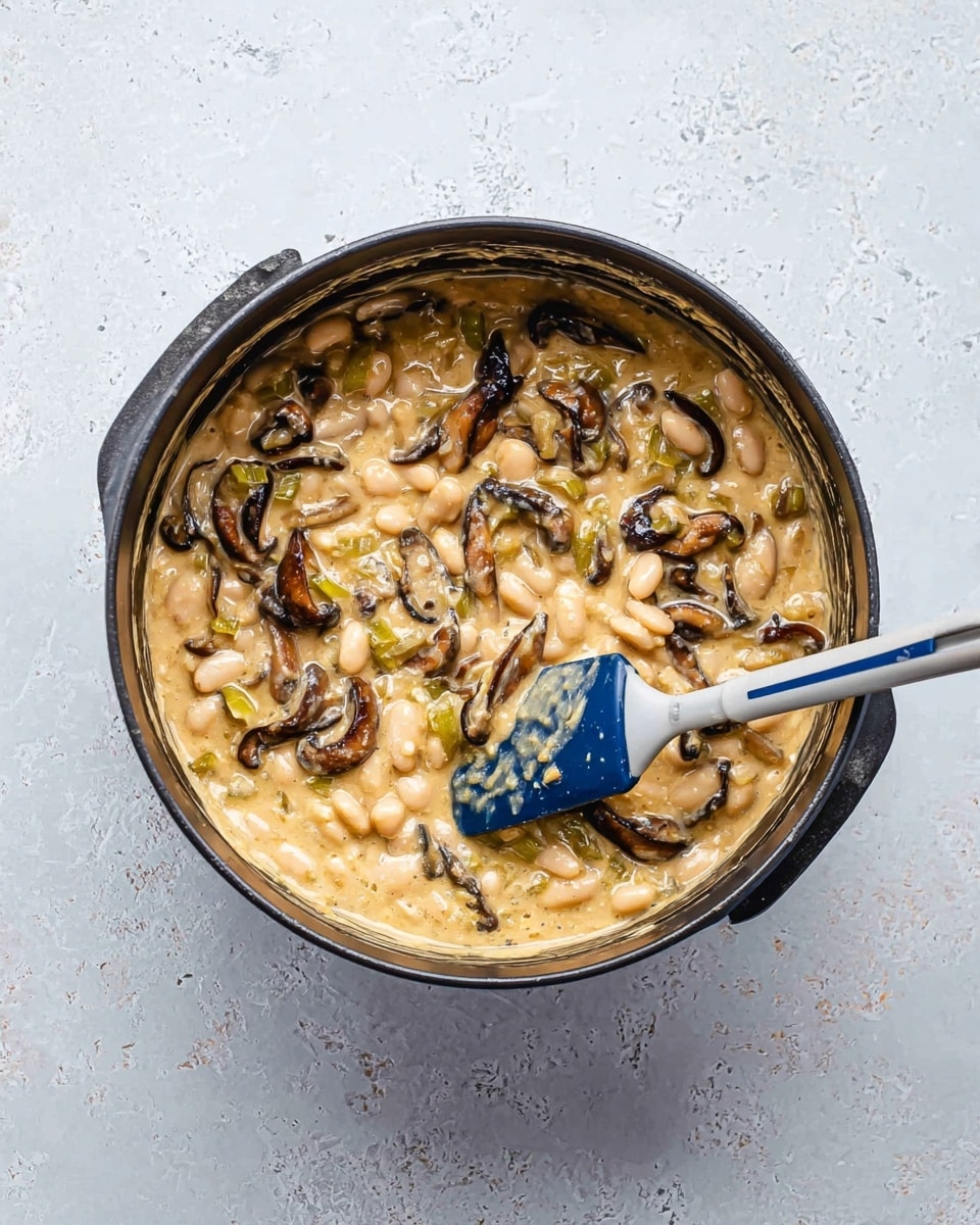 A round black pot filled with a creamy mixture with three main layers: white beans, dark brown sliced mushrooms, and small pieces of light green vegetables, all mixed together in a thick beige sauce with a slightly shiny texture. A blue and white spatula is resting inside the pot, slightly lifted with some sauce on it. The pot is placed on a white marbled textured surface. photo taken with an iphone --ar 4:5 --v 7