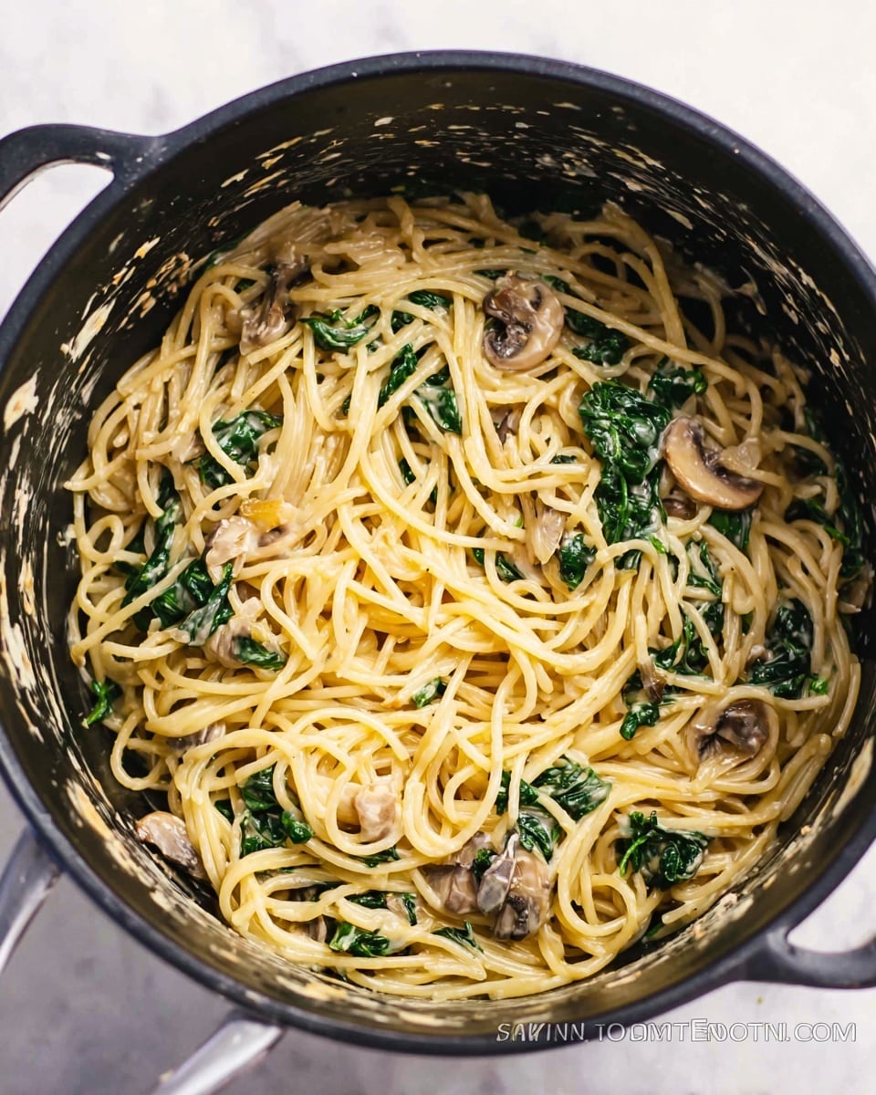 Inside a black cooking pot, there is one main layer of spaghetti pasta mixed with creamy sauce that lightly coats the noodles. Mixed within the spaghetti are pieces of cooked dark green spinach and small slices of brown mushrooms. The creamy texture is smooth and light, blending with the yellowish pasta and contrasting with the dark greens and browns. The pot edges show some sauce splatter, adding a casual homemade feel. The whole scene is set on a white marbled surface. photo taken with an iphone --ar 4:5 --v 7