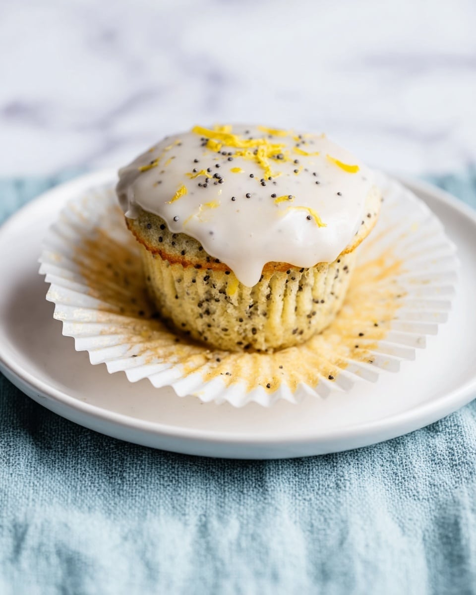 A single poppy seed muffin is shown with its white paper wrapper peeled back and spread out on a white plate. The muffin has a light yellow color with black poppy seeds visible throughout. On top, there is a smooth, creamy white icing with some small black poppy seeds and tiny yellow zest pieces sprinkled over it. The plate sits on a soft, light blue textured cloth, all set against a white marbled surface. photo taken with an iphone --ar 4:5 --v 7