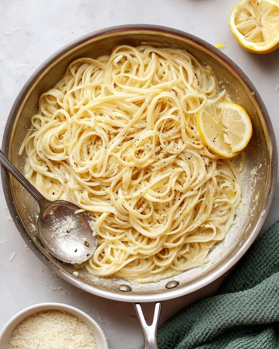 A silver pan filled with light yellow creamy pasta noodles piled in the center, with a single lemon wedge resting on top on the right side. The pasta is coated with a smooth sauce, sprinkled with small bits of black pepper and tiny lemon zest pieces. A large silver spoon sits partially under the pasta on the left side of the pan. The pan is on a white marbled surface. Below the pan, there is a small white bowl filled with grated cheese, and on the right side is a green cloth. Photo taken with an iphone --ar 4:5 --v 7
