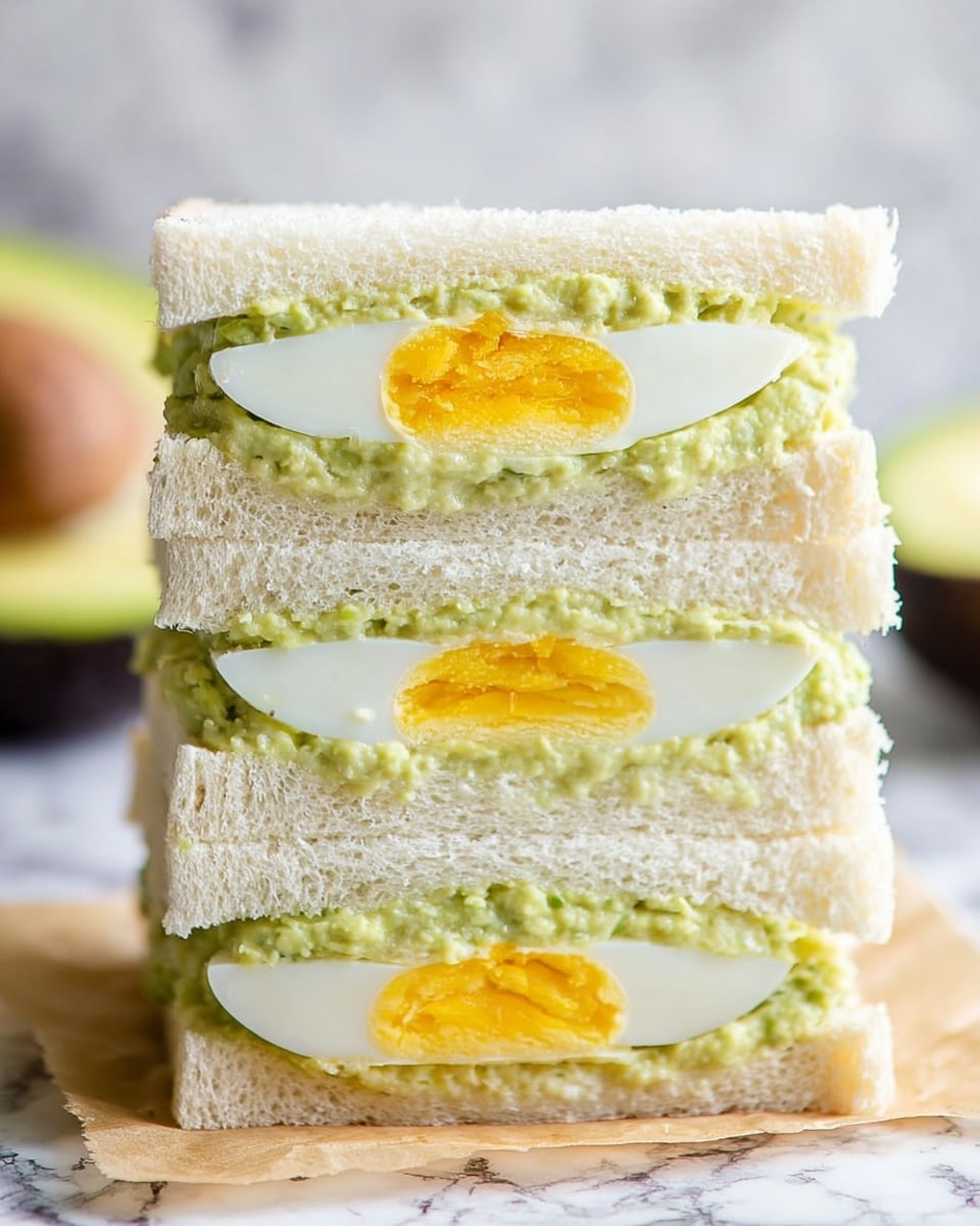 The image shows a stack of three sandwiched layers of soft, white bread with a light, airy texture. Between each bread layer, there is a thick spread of creamy, pale green avocado mash that looks slightly chunky. In the center of each layer, a halved boiled egg with a firm white outer layer and a bright yellow yolk is visible, positioned horizontally so the yolk faces the front. The sandwich is placed on a piece of parchment paper on a surface with a white marbled texture. In the background, you can see part of a halved avocado, adding a touch of dark green color. Photo taken with an iphone --ar 4:5 --v 7