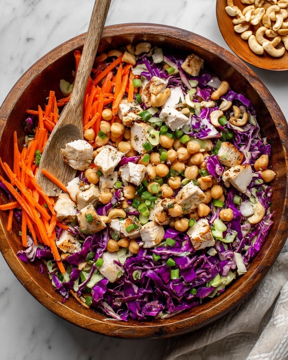 The image shows a large wooden bowl filled with a colorful salad made of multiple layers. The bottom layer has bright purple cabbage chopped into small pieces, with thin, orange carrot sticks spread evenly on top. Scattered throughout the salad are chunks of grilled white chicken with a light brown sear, creamy beige chickpeas, and whole cashew nuts. The salad is topped with finely chopped green onions, adding fresh green color. A long wooden spoon is placed inside the bowl, resting on the left side. The bowl is set on a white marbled surface, and there is a small wooden plate with more cashews in the upper right corner of the image. photo taken with an iphone --ar 4:5 --v 7