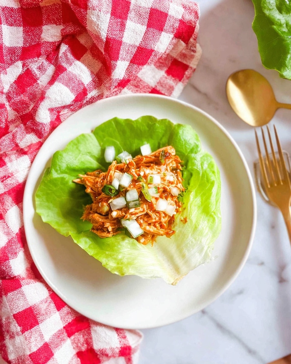 A single green lettuce leaf is placed in the center of a white plate, forming the base layer. On top of the lettuce leaf, there is a layer of shredded orange-brown chicken mixed with a sauce. The chicken layer is topped with small white pieces of chopped onions, scattered unevenly. The plate is on a white marbled surface with a red and white checkered cloth and a golden spoon partly visible in the background. Photo taken with an iphone --ar 4:5 --v 7