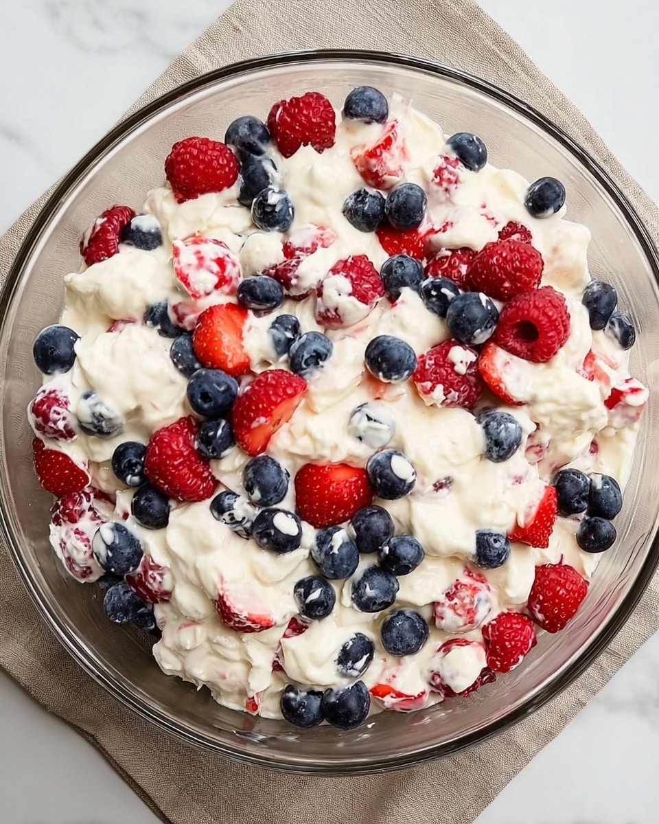 A clear glass bowl filled with a creamy white mixture with uneven dollops throughout, containing fresh blueberries, red raspberries, and halved strawberries mixed evenly within the creamy layer, giving a colorful contrast of deep blue, bright red, and white. The bowl sits on a neutral beige cloth over a white marbled surface. The mixture looks soft and fluffy with the berries partially embedded and resting on top. photo taken with an iphone --ar 4:5 --v 7
