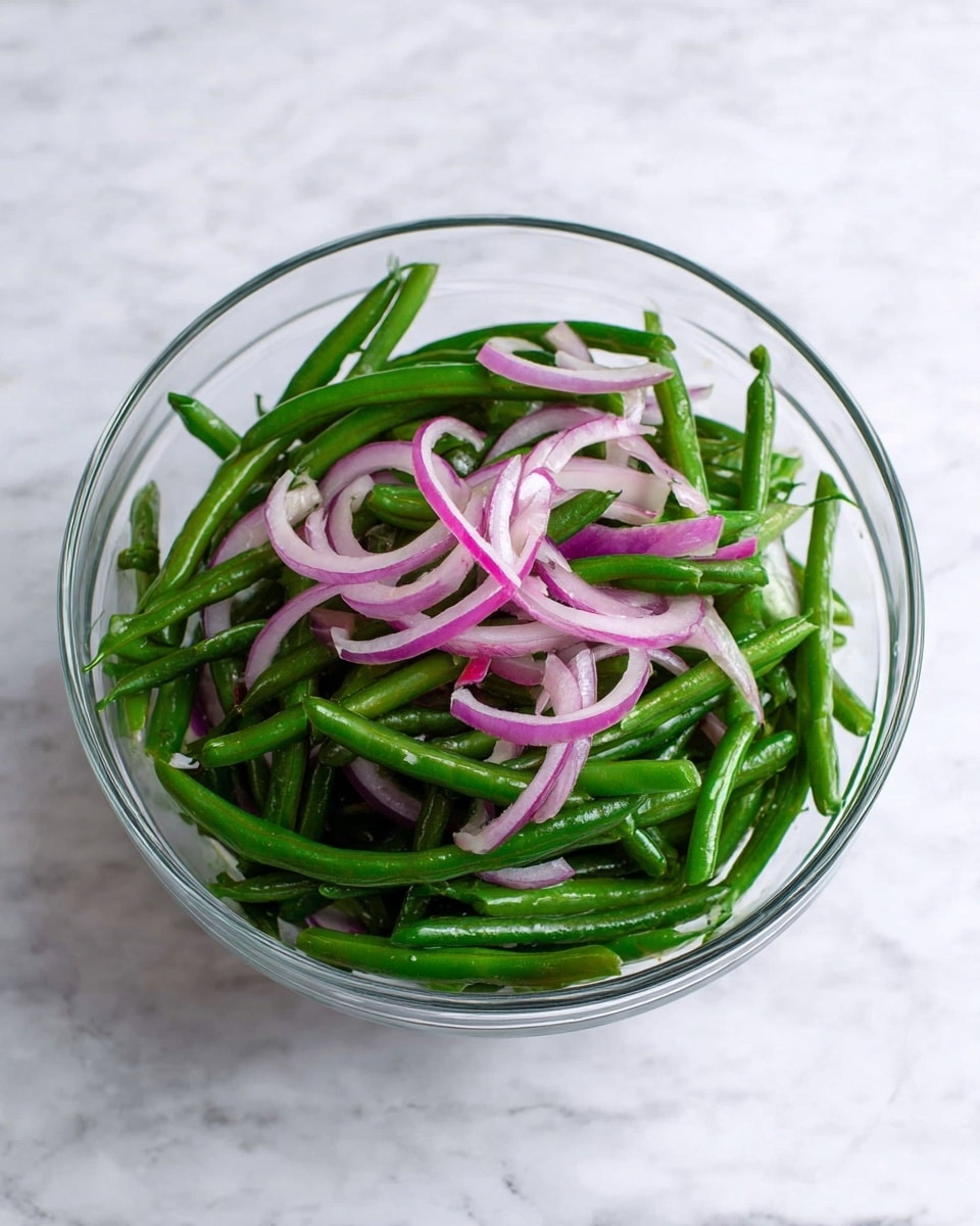 A clear glass bowl holds a fresh green bean salad with two main layers. The bottom layer is made up of bright green, whole green beans that are crisp and shiny. The top layer consists of thin curved slices of purple-red onion, adding a slight contrast in color and texture. The bowl is placed on a surface with a white marbled texture. photo taken with an iphone --ar 4:5 --v 7