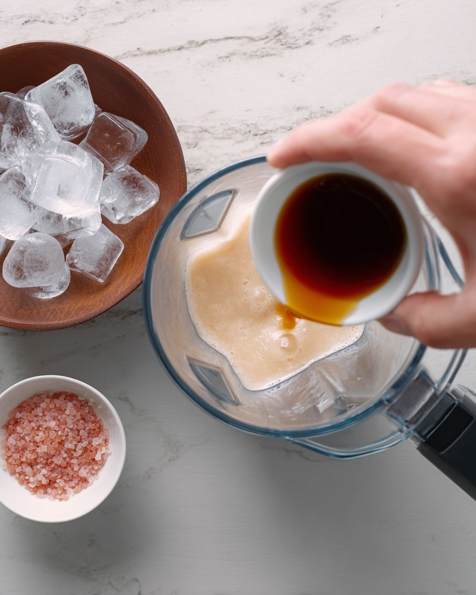 A woman's hand is pouring a small amount of dark brown liquid from a small white dish into a clear blender container. Inside the blender, there is a light beige foamy liquid layer. To the left of the blender is a brown bowl filled with clear ice cubes, and to the right is a small white bowl with pink coarse salt. The surface under all items is white marbled texture. photo taken with an iphone --ar 4:5 --v 7