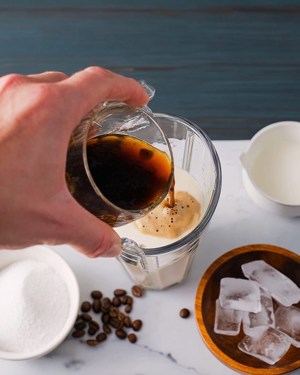 A woman's hand is pouring dark brown coffee with light froth from a small clear glass into a tall clear blender container filled with white milk, creating a mixing effect visible through the glass. To the right side, there is a white bowl with white granulated sugar and a wooden bowl containing clear ice cubes. Coffee beans are scattered on a white marbled surface around the bowls. Photo taken with an iphone --ar 4:5 --v 7