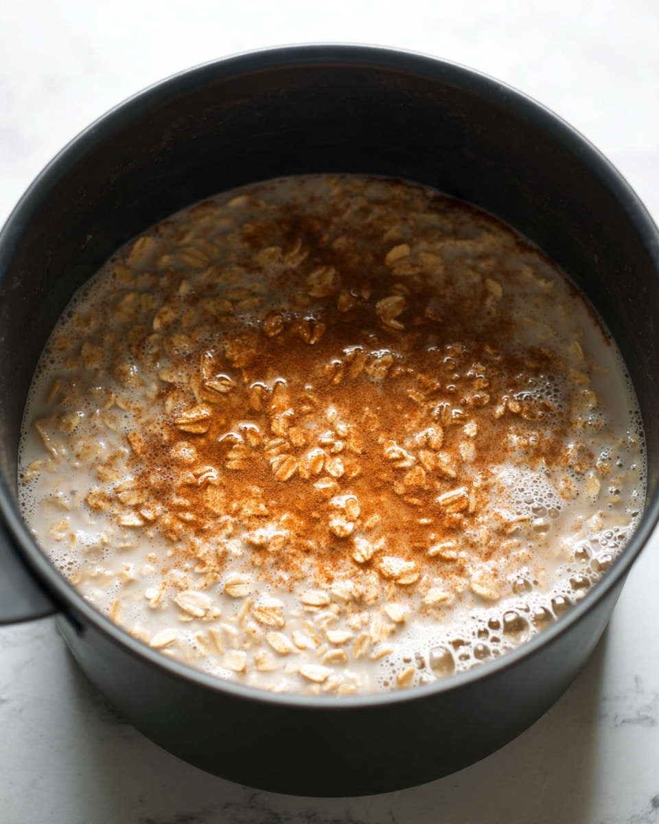 The image shows a black pot filled with oatmeal cooking in milk, with visible light brown oat flakes floating on the surface. A layer of cinnamon powder with a warm reddish-brown color is sprinkled on top of the oats in a somewhat uneven pattern, mostly concentrated toward one side. The oatmeal mixture looks creamy with some bubbles around the edges. The pot is set on a white marbled surface. Photo taken with an iphone --ar 4:5 --v 7