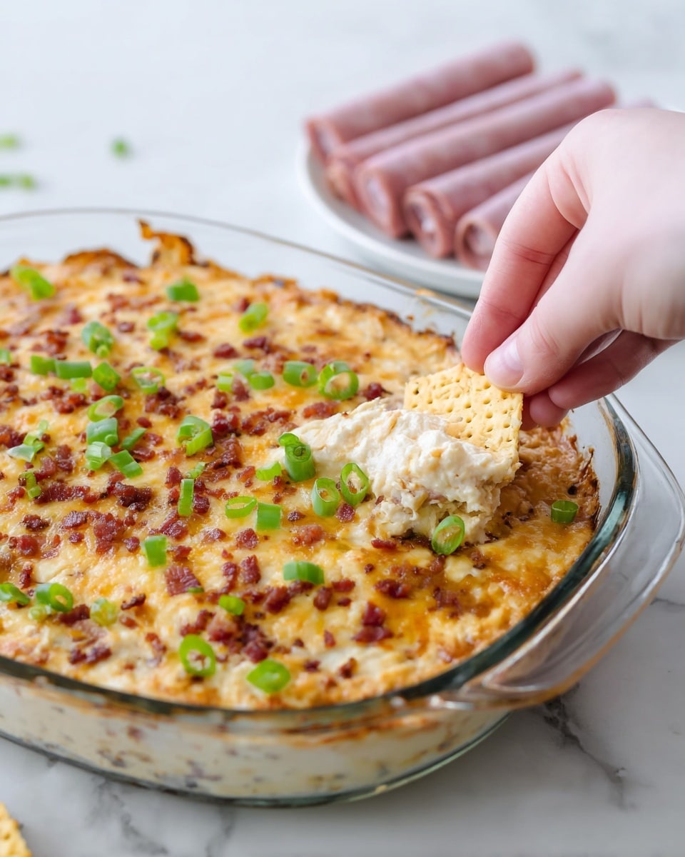 A glass baking dish filled with a thick, creamy cheese dip that has a golden-brown browned top layer mixed with small reddish bits, possibly bacon or seasoning. The dip is garnished with chopped green onions scattered on the surface. A woman's hand is dipping a light brown woven-texture cracker into the cheese dip near the corner of the dish. The background and surface have a white marbled texture, and there is a white plate with rolled slices of pink deli meat slightly blurred in the background. Photo taken with an iphone --ar 4:5 --v 7