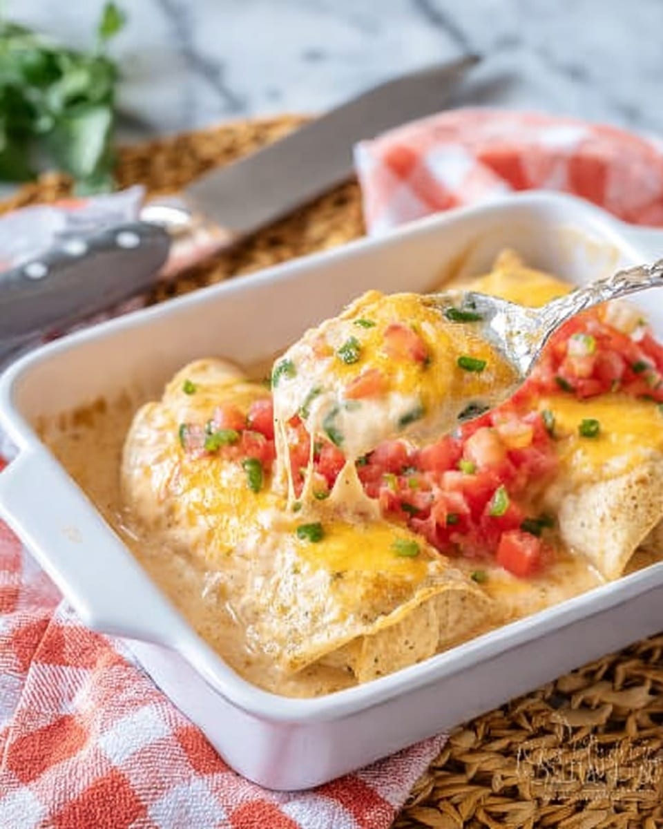 The dish is served in a white rectangular baking dish filled with three soft, folded layers of light golden tortillas covered in creamy, light brown sauce. On top, there is melted yellow cheese with bits of diced red tomatoes and green peppers sprinkled over it. A spoon lifts one of the folded tortillas, showing a smooth sauce dripping slightly. The dish sits on a woven mat with a white marbled surface visible around it. A silver knife and a folded red and white checkered napkin are beside the dish. Photo taken with an iphone --ar 4:5 --v 7