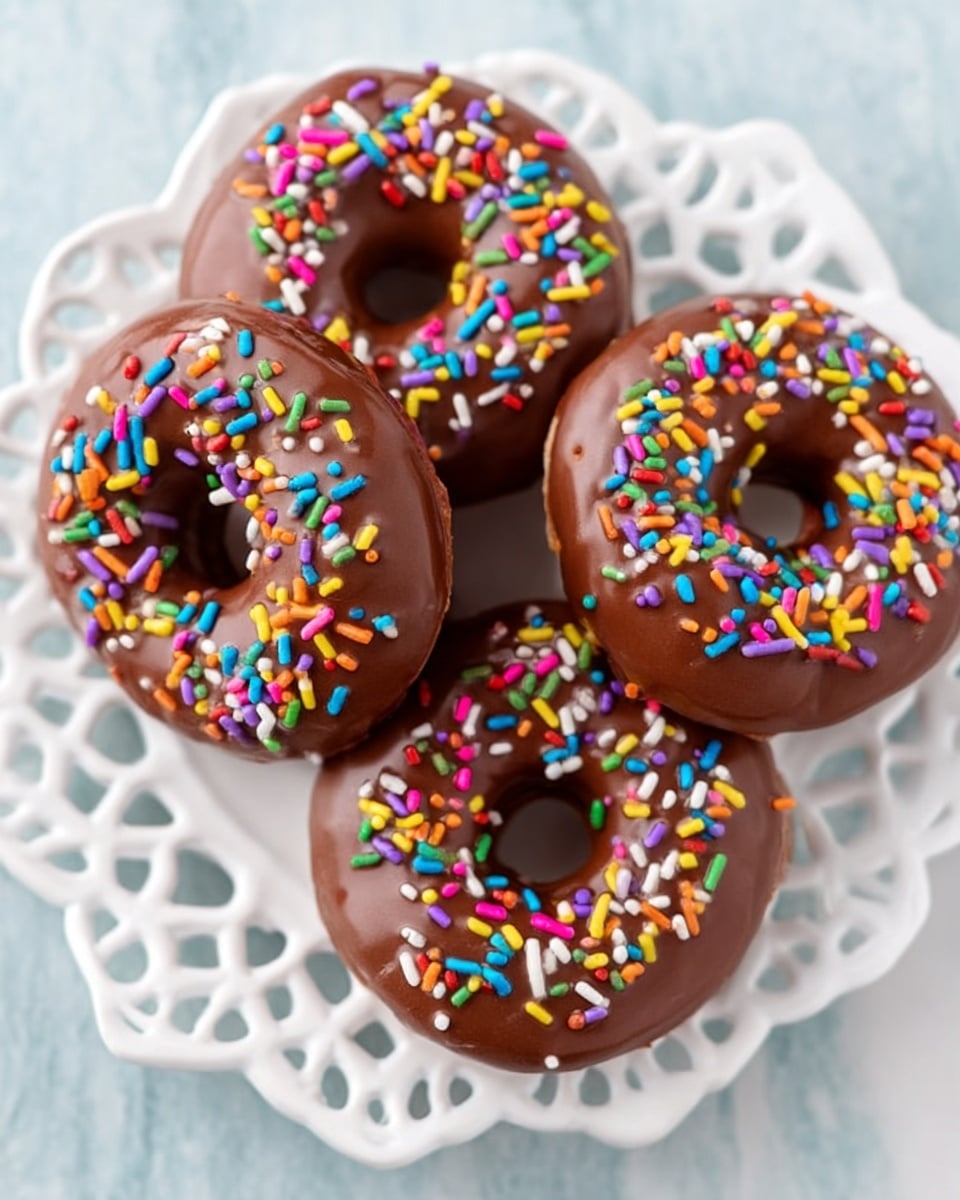 Four round donuts are placed closely together on a white decorative plate with cutout patterns along the edge. Each donut has a smooth milk chocolate glaze layer covering the top, giving a shiny texture. On top of the chocolate glaze, there is a colorful mix of small cylindrical sprinkles in red, yellow, green, purple, blue, orange, pink, and white spread evenly. The plate sits on a white marbled textured surface with soft light shining from above, capturing the vibrant colors and glossy finish of the donuts. photo taken with an iphone --ar 4:5 --v 7