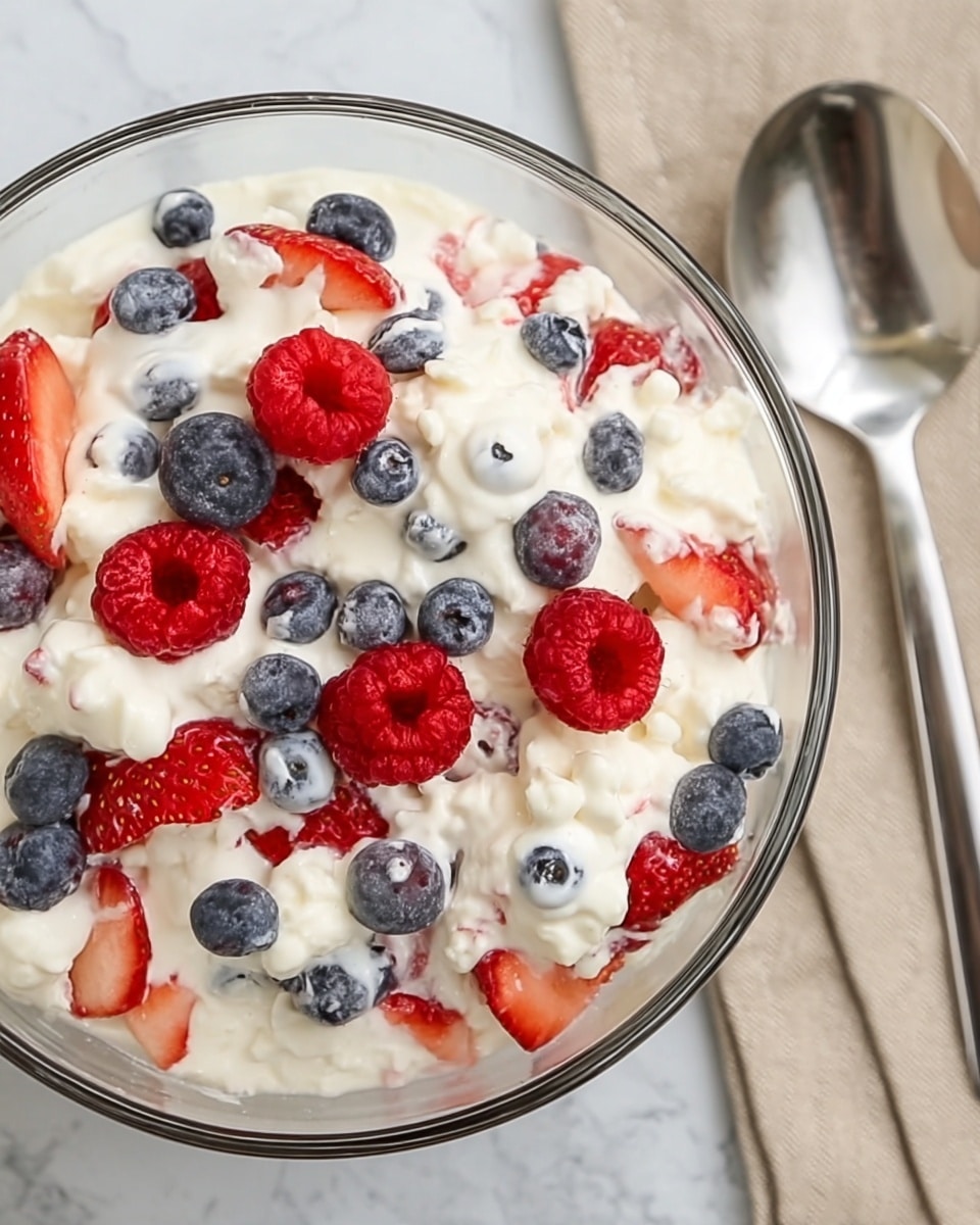 A clear glass bowl filled with a creamy white mixture that has a fluffy texture, with fresh red strawberries sliced in half, whole red raspberries, and small round blue blueberries scattered evenly throughout. The mixture looks thick and covers the fruits partially, showing red and blue colors popping against the white cream. The bowl sits on a white marbled surface with a silver spoon placed to the right on a beige cloth. photo taken with an iphone --ar 4:5 --v 7