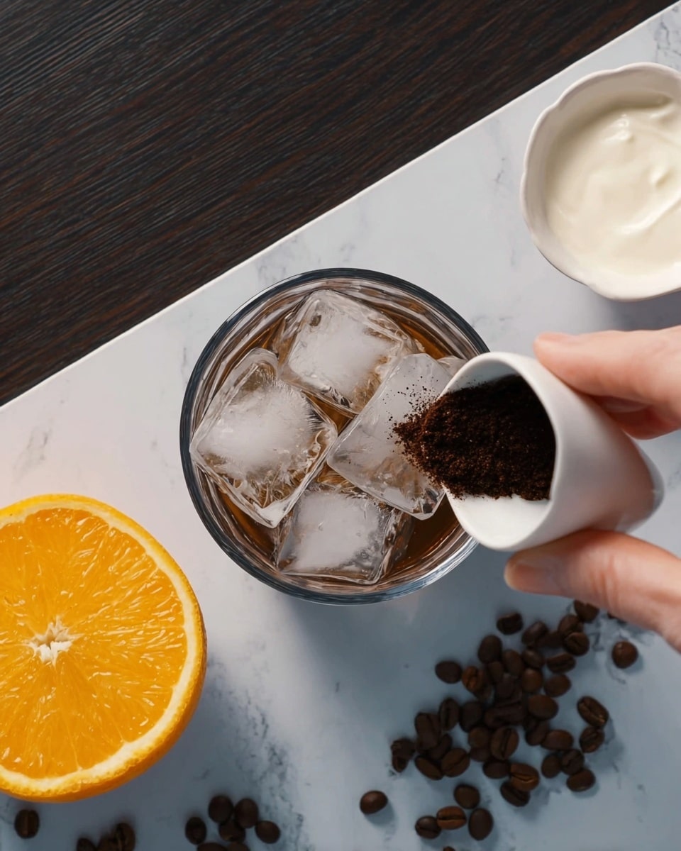 A clear glass filled with several large ice cubes sits at the center on a white marbled surface. A woman's hand is pouring dark brown coffee grounds from a small white bowl into the glass, adding texture to the ice. To the left of the glass is a halved orange showing its bright, juicy orange segments. On the upper right, there is a small white bowl filled with white cream or milk. Scattered coffee beans surround the bowl, adding a rich brown contrast against the white marbled surface. Photo taken with an iphone --ar 4:5 --v 7