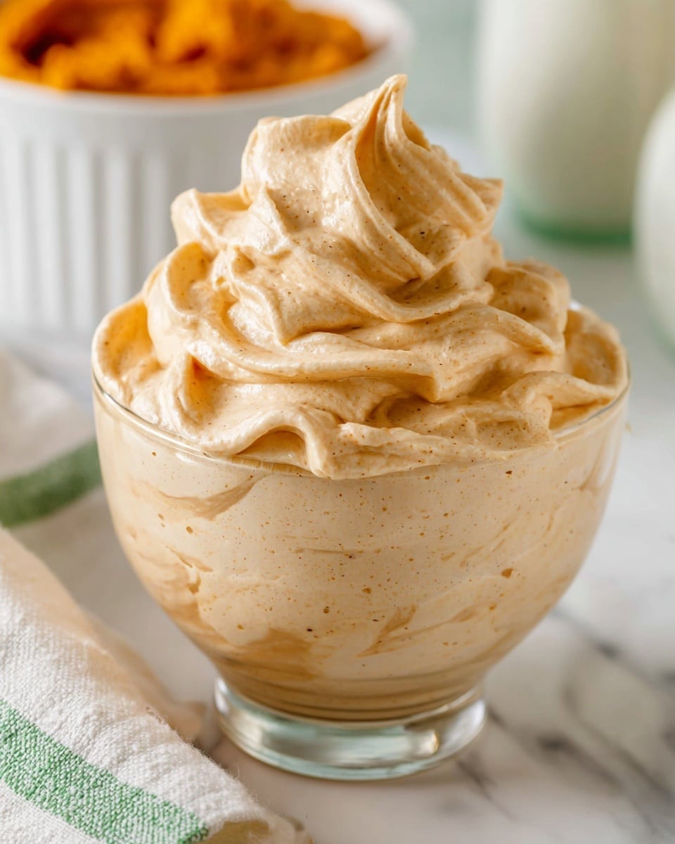 A clear glass bowl filled with thick, creamy light brown whipped cream with visible tiny darker specks, swirled in a tall, ruffled pattern that rises above the bowl's top edge, showing smooth, soft texture with gentle folds. The bowl sits on a white marbled surface beside a white cloth with a green stripe, and a blurred white bowl filled with an orange mixture is in the background. Photo taken with an iphone --ar 4:5 --v 7