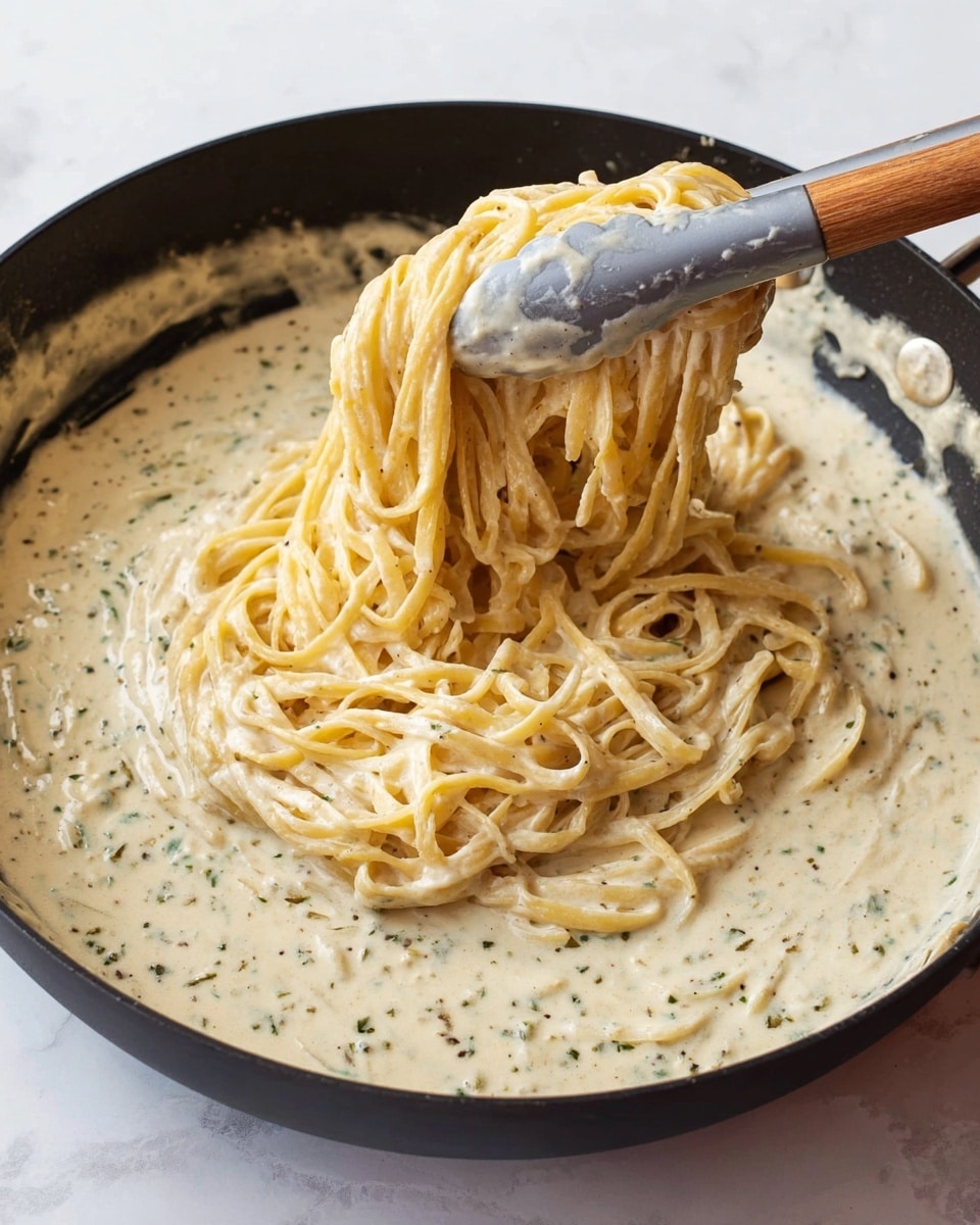 A black pan filled with creamy white sauce mixed with herbs that give small green and black specks throughout. In the middle, a layer of yellowish pasta coated thickly with the creamy sauce is being lifted by gray kitchen tongs with a wooden handle from the right side. The texture of the sauce is smooth and rich, covering the pasta evenly. The pan sits on a white marbled surface. Photo taken with an iphone --ar 4:5 --v 7