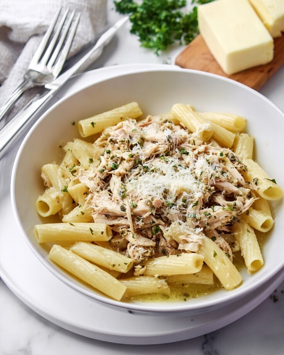 A white bowl filled with two main layers: at the bottom, cooked penne pasta in pale yellow with smooth surfaces and ridges, and on top, shredded light brown chicken mixed with small green herb flakes, sprinkled with finely grated white cheese creating a soft texture over the dish. The bowl is placed on a white marbled surface with a fork and knife nearby, and a block of butter and some green herbs are slightly visible in the background. Photo taken with an iphone --ar 4:5 --v 7