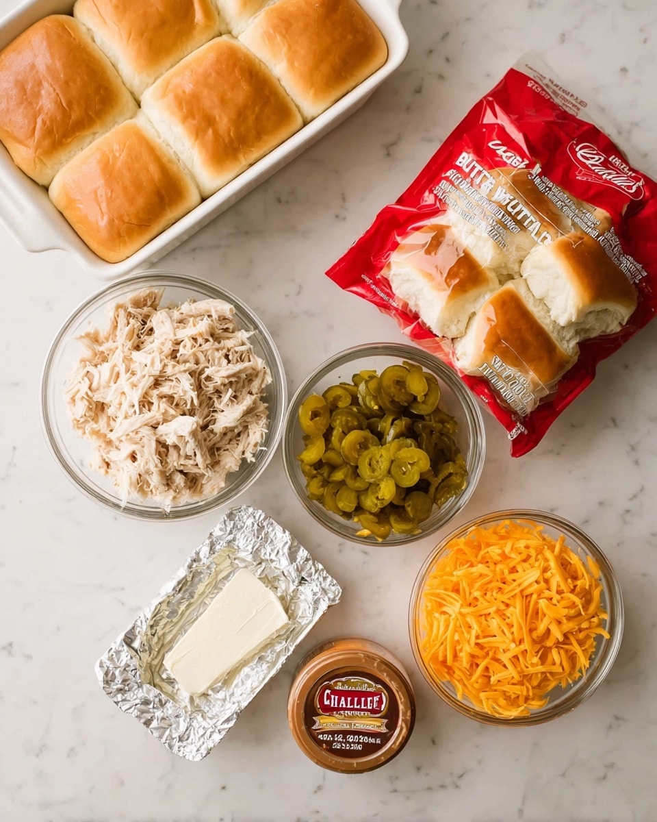 The image shows ingredients arranged on a white marbled surface for making sliders. There is a clear bowl filled with shredded light beige cooked chicken. Next to it is a smaller clear bowl with sliced yellow-green pickled jalapeños. Another clear bowl holds bright orange shredded cheddar cheese. A rectangular silver foil wrapper is open, showing a white creamy spread inside. Below this is a small brown and red container of seasoned butter spread labeled