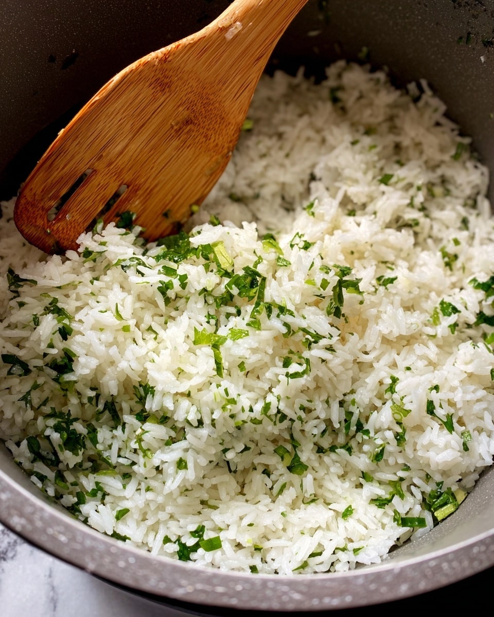 A close-up view of cooked white rice mixed with small, bright green herb pieces inside a gray cooking pot, with a wooden spatula resting on the rice toward the top left. The rice grains are separate, soft-textured, and spread evenly across the bottom layer of the pot. The gray pot edge frames the rice lightly, and a hint of a white marbled surface is visible around the base of the pot. photo taken with an iphone --ar 4:5 --v 7