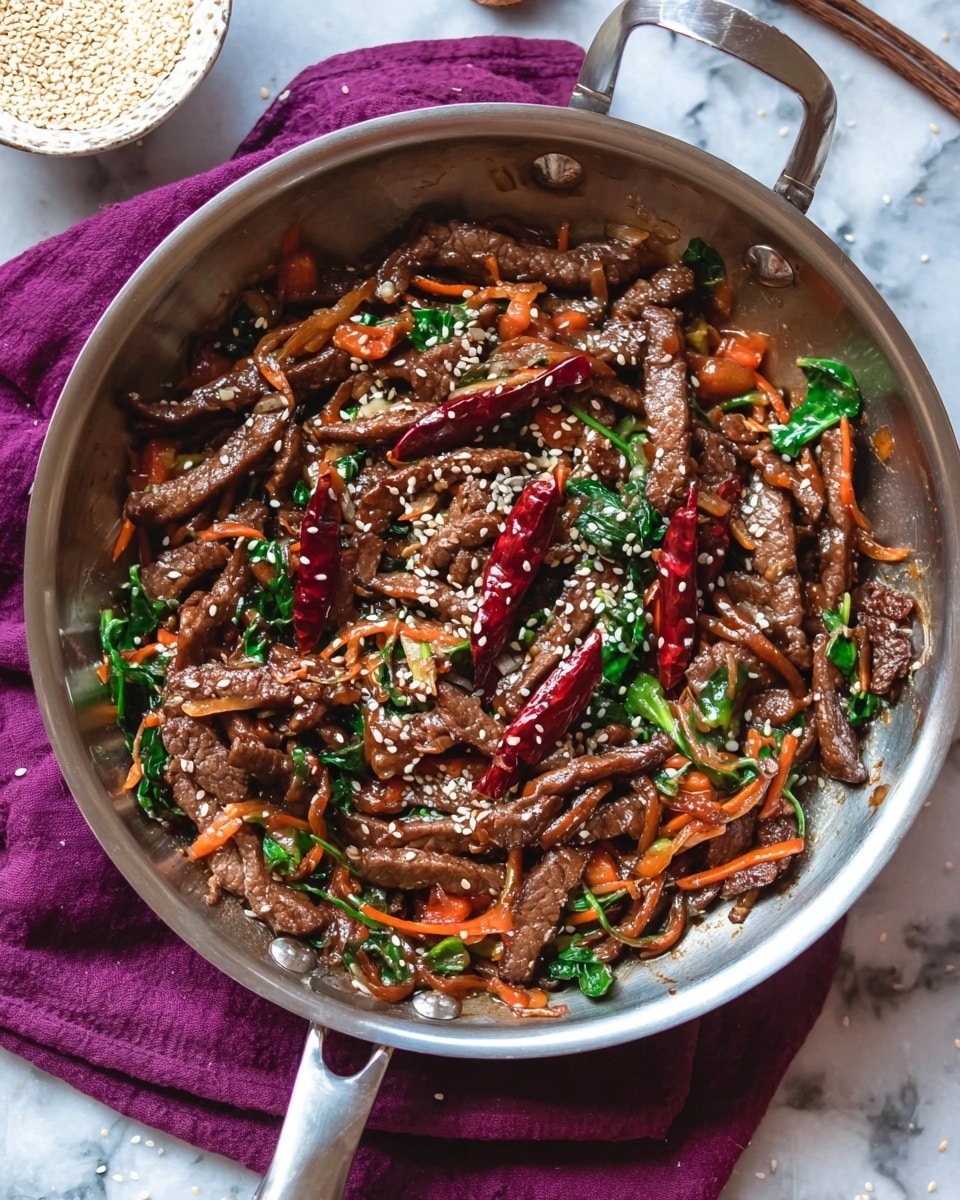 The image shows a metal pan filled with a stir-fry dish. The dish has many thin, brown strips of cooked meat layered with small orange carrot pieces and green leafy vegetables scattered throughout. There are also several whole dried red chili peppers placed on top and mixed inside. The dish is sprinkled with white sesame seeds, adding texture and contrast. The pan is placed on a folded purple cloth on a white marbled surface, and a small bowl with light sesame seeds is visible in the background. photo taken with an iphone --ar 4:5 --v 7