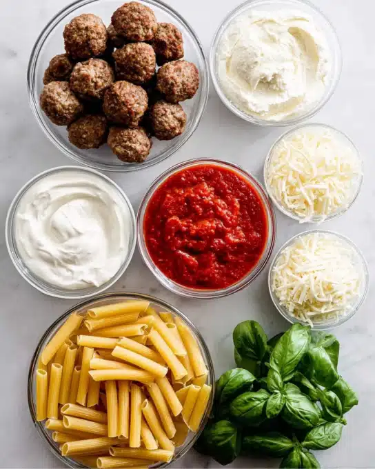 The image shows seven clear bowls arranged on a white marbled surface, each with different ingredients. The largest bowl at the bottom holds dry yellow pasta, tube-shaped and smooth. Above it to the left is a bowl filled with several cooked brown meatballs with a slightly rough texture. Next to the meatballs on the left is a white bowl with thick and creamy white Greek yogurt, smooth and swirled. Above the pasta in the middle is a bowl of bright red marinara sauce with a chunky texture. To the right of the marinara sauce is a smaller bowl containing shredded white mozzarella cheese with a soft texture. Above the mozzarella is another tiny bowl with finely grated white parmesan cheese, fluffy and light. Next to all the bowls on the right side is a bunch of fresh green basil leaves with a smooth and slightly shiny surface. Photo taken with an iphone --ar 4:5 --v 7