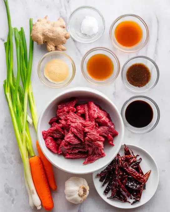The image shows a top view of fresh ingredients for Sha Cha Beef Stir Fry arranged neatly on a white marbled surface. In the center, there is one white bowl filled with bright red strips of raw beef, with a rough texture. Surrounding the bowl are multiple small white and clear glass bowls holding different sauces and pastes, varying in color from dark brown, light tan, reddish-brown, to black. To the left, there are fresh green onions with vibrant green stalks and white roots, next to a bright orange carrot and a whole head of garlic with a rough textured peel. Below the beef bowl, a small white plate holds several dried red chili peppers, wrinkled and dark red. The setup is clean and well-organized, with all ingredients clearly visible and spaced evenly. photo taken with an iphone --ar 4:5 --v 7