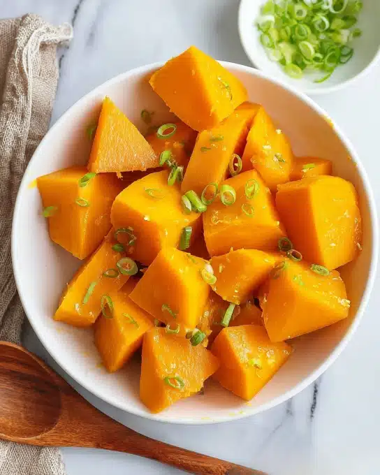 A white bowl filled with several chunks of bright orange cooked pumpkin, each piece showing a smooth yet slightly soft texture, topped with small, scattered pieces of chopped green onion. The bowl sits on a white marbled surface next to a small white bowl holding more chopped green onions and a wooden spoon. A small green plant is visible on the upper left corner. The overall colors are warm and fresh with the bright orange pumpkin contrasting with the green onions and white bowls photo taken with an iphone --ar 4:5 --v 7