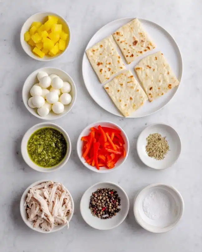 Parmesan Garlic Chicken Pizza Recipe 4 A white plate holds a flat bread folded into four sections at the top of the image. Below it, six small white bowls are arranged in a grid pattern on a white marbled surface. The top left bowl contains bright yellow pineapple pieces, next to it the top middle bowl has small white fresh mozzarella balls, and to the top right a bowl filled with thin red bell pepper strips. In the middle row on the left, there is a bowl of green pesto sauce, to the right, a bowl with small black peppercorns. The bottom row on the left holds shredded light brown rotisserie chicken, and to the right is a bowl with white salt. The colors contrast brightly against the clean white marbled surface. Photo taken with an iphone --ar 4:5 --v 7