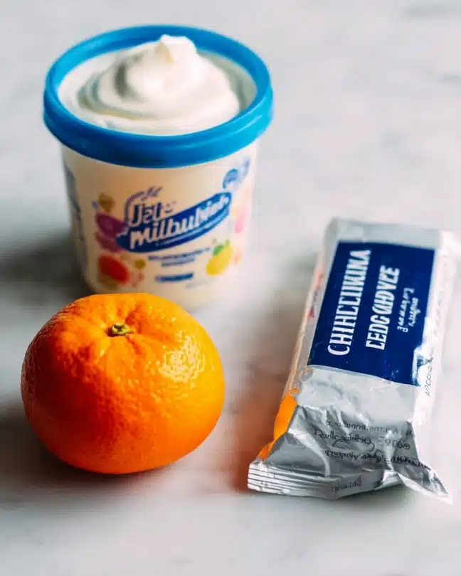 Fruit Rainbow Platter with Cream Cheese-Marshmallow Dip Recipe 4 The image shows a container of Jet-Puffed marshmallow creme with a white jar and a blue lid on the left side, a bright orange clementine fruit in the middle, and a rectangular package of Philadelphia original cream cheese with a silver wrapper and dark blue text on the right. All items are placed on a white marbled texture surface with a soft, neutral background. photo taken with an iphone --ar 4:5 --v 7