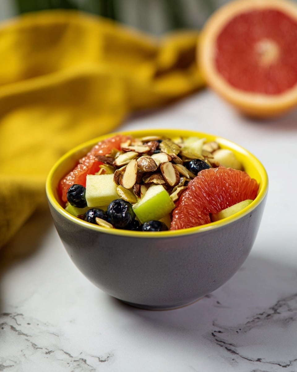 A short gray bowl with a yellow inside is full of a fruit mix. The bottom layer shows green apple pieces and dark blue blueberries evenly spread. On top of that is a layer of red-orange grapefruit wedges with some shine. The top layer is scattered chopped almonds, light brown with rough edges. The bowl sits on a white marbled surface with a blurred yellow cloth to the left and a half of a red grapefruit to the right in the background. photo taken with an iphone --ar 4:5 --v 7