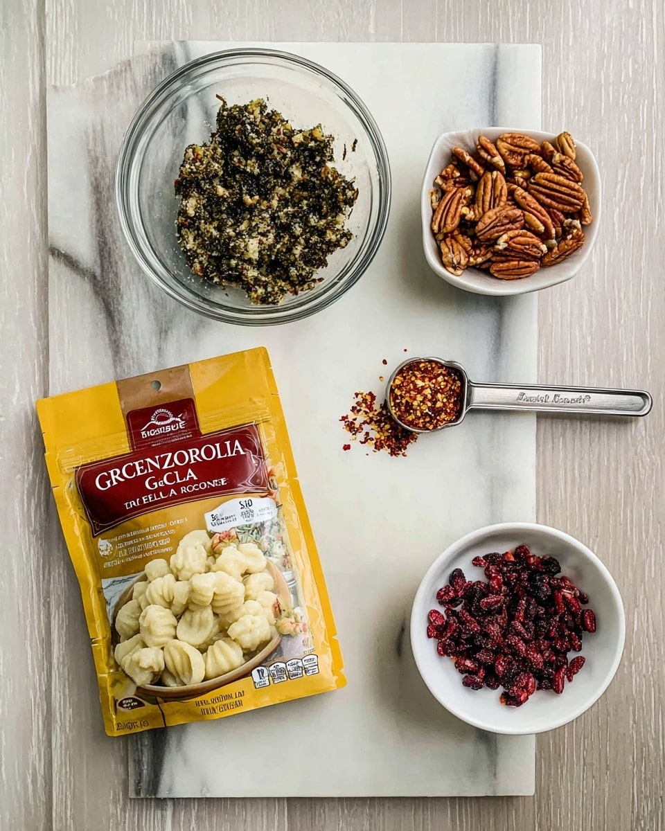 The image shows five items arranged on a white marbled surface: a clear glass bowl filled with a dark green chunky mixture, a small metal measuring spoon holding red pepper flakes, a white bowl with pecan nuts, a clear glass bowl with small dried red berries, and a yellow package labeled