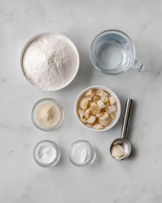 Vegan Marshmallow Recipe 4 The image shows seven containers with different ingredients arranged on a white marbled surface. At the top left, there is a large white bowl filled with fine white sugar. To its right, a clear glass mug holds water. Below the water, a small clear glass bowl contains a light beige powder. In the center, a white bowl is filled with light brown, crumbly chunks. To the left of this bowl, a tiny clear glass bowl has a white powdery ingredient. Below, two small clear bowls contain white powdered ingredients with different textures. To the right of these, a metal measuring spoon with a white powder is placed flat on the surface. The text