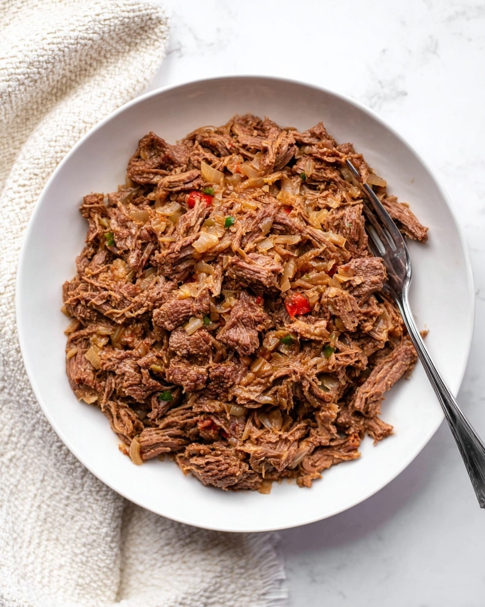 This image shows a white round plate filled with shredded brown cooked beef mixed with bits of cooked onions and some small red and green pieces, likely peppers or seasoning. The beef appears moist and tender with uneven chunks and shreds piled together in a single thick layer covering the plate surface. A silver fork is placed on the right side of the plate, partially resting on the beef. The plate sits on a white marbled surface with a cream-colored textured cloth partly visible in the top left corner. photo taken with an iphone --ar 4:5 --v 7