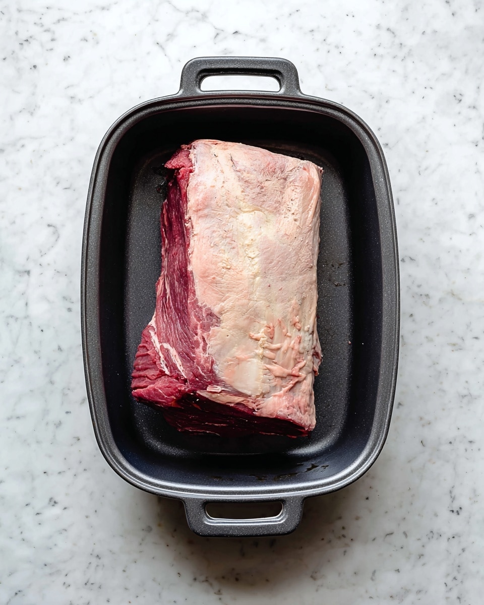 A large piece of raw meat with a thick layer of pale fat on top and red muscle underneath sits in a dark gray cooking pan with handles on both ends, placed on a white marbled surface. The meat is rectangular and fills the pan almost fully, showing rough texture and natural color shades of raw beef. Photo taken with an iphone --ar 4:5 --v 7