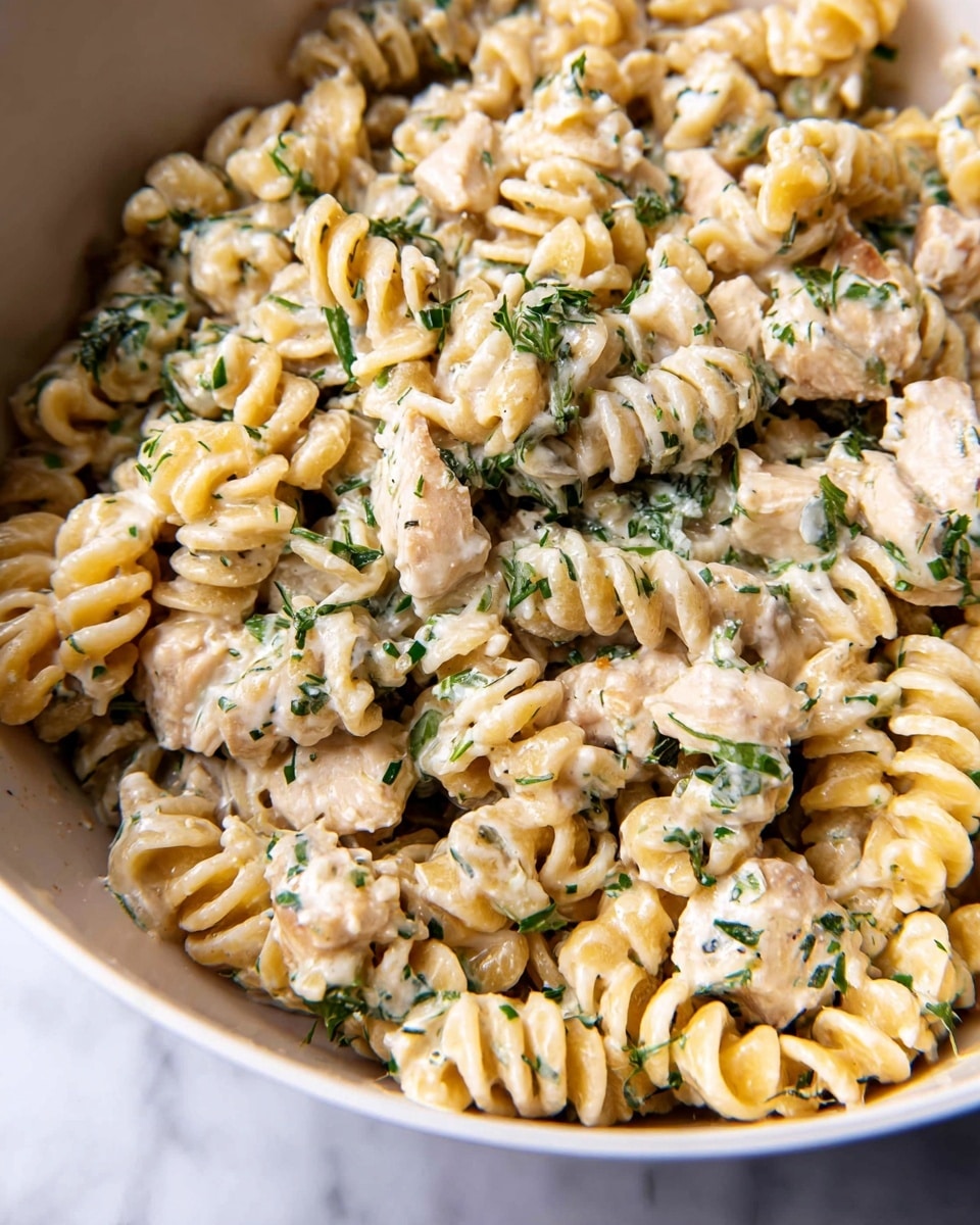 The dish shows a close-up view of small, curly pasta in a creamy sauce mixed with finely chopped green herbs and pieces of light-colored chicken. The pasta is pale yellow and looks soft, while the sauce is thick and white, coating the pasta and chicken evenly. The green herb bits are spread throughout, adding small bursts of color. The food is in a white bowl with a smooth edge, and the background shows a clean white marbled surface. photo taken with an iphone --ar 4:5 --v 7