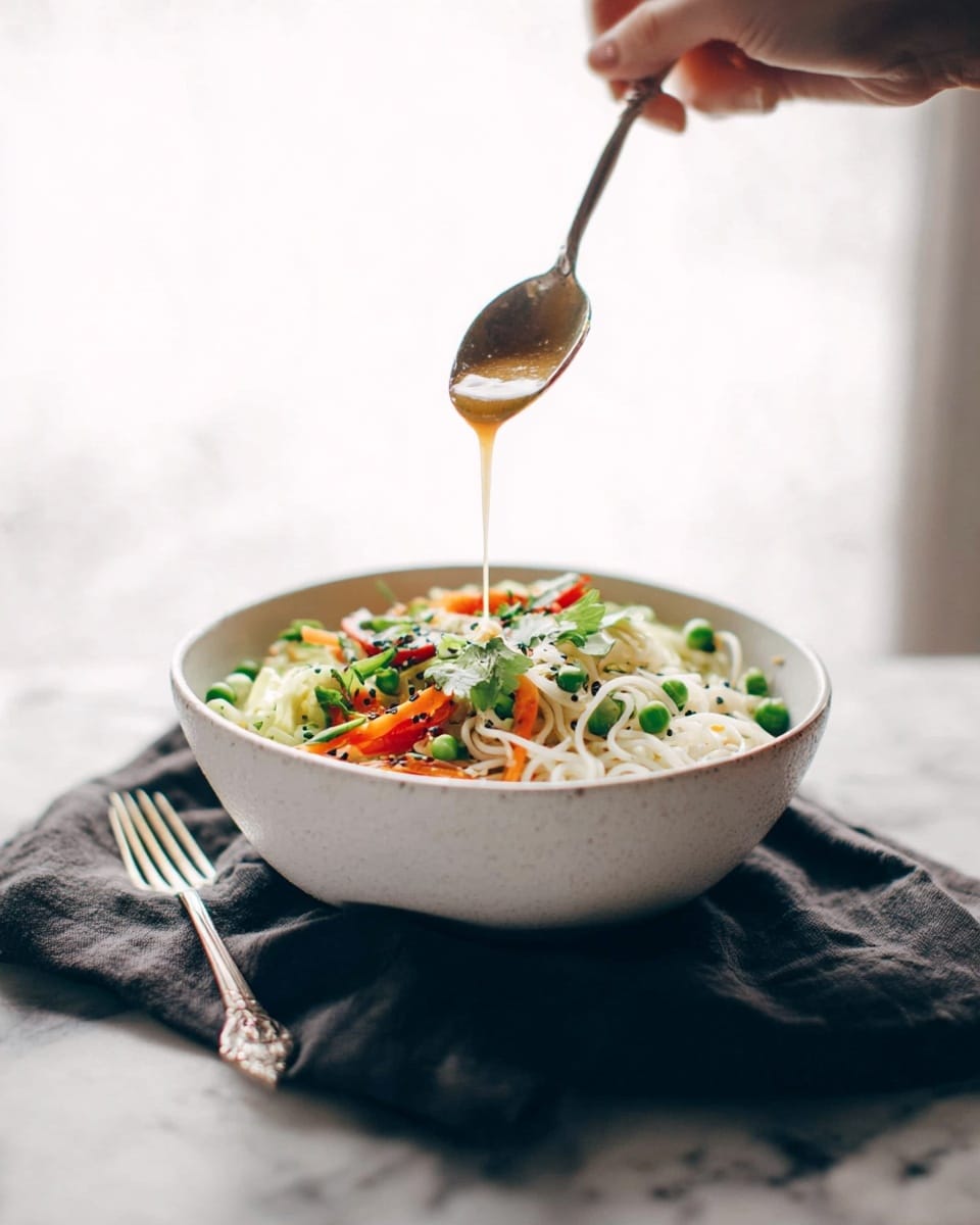 A white bowl is filled with a colorful noodle dish with visible layers of white noodles at the bottom, bright green peas and herbs scattered on top, orange carrot strips, and red bell pepper pieces mixed in. A woman's hand holds a spoon above the bowl, pouring a light brown sauce over the noodles. The bowl sits on a dark cloth on a white marbled surface, with a silver fork placed on the left side of the bowl. The background is softly lit and blurry, making the bowl and the woman's hand the main focus. photo taken with an iphone --ar 4:5 --v 7