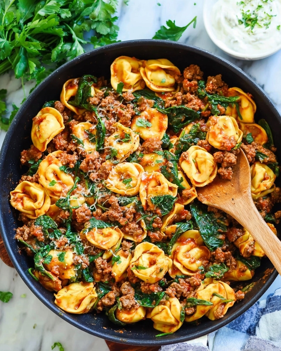 The image shows a black pan filled with tortellini pasta mixed with ground meat and spinach. The pasta is yellow with a smooth texture and is coated in a reddish sauce. The ground meat is brown and crumbly, spread evenly throughout the dish. Bright green spinach leaves add a fresh contrast, scattered all around. A wooden spoon lifts a portion from the pan, showing the mix of pasta, meat, and spinach. The pan sits on a white marbled surface with some parsley leaves nearby and a small white bowl with a white creamy dip in the background. Photo taken with an iphone --ar 4:5 --v 7