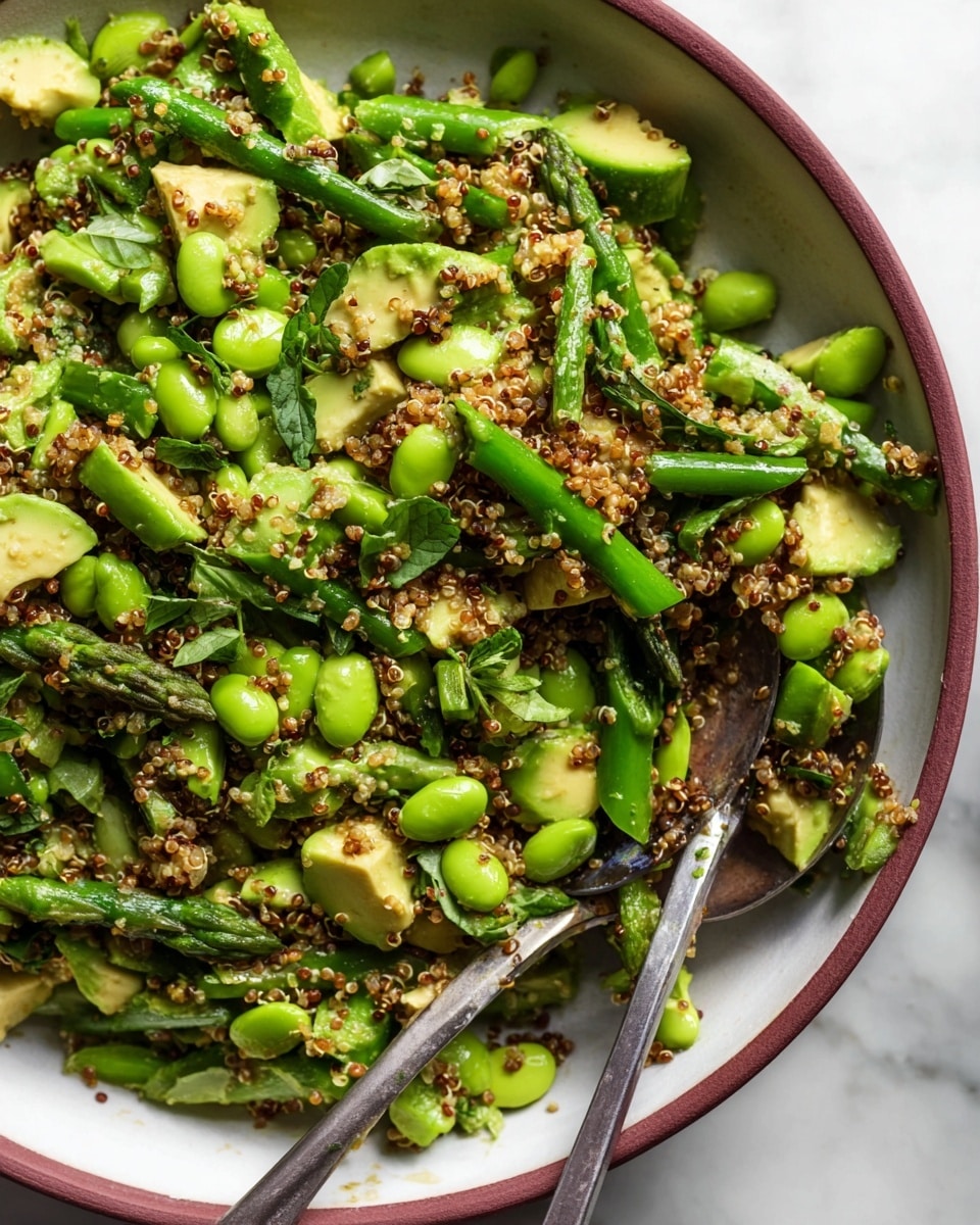 The image shows a close-up of a salad in a white bowl filled with bright green asparagus pieces, green edamame beans, chunks of avocado, and cooked brown quinoa. The asparagus is cut into medium-sized pieces scattered throughout, while the avocado pieces are soft and slightly creamy with a light green color. The quinoa is fluffy and adds small, round, textured grains that coat the vegetables lightly. The salad has some green leafy herbs mixed in, adding depth to the green colors. A metal serving spoon and fork rest in the bowl, partially covered by the salad. The bowl sits on a white marbled surface. photo taken with an iphone --ar 4:5 --v 7