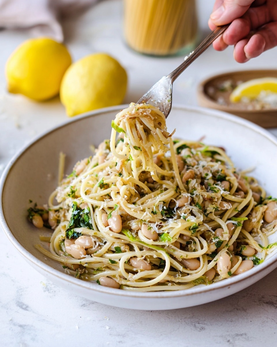 A white bowl on a white marbled surface holds a large serving of spaghetti mixed with small white beans and finely chopped greens. The pasta is light yellow with a slightly glossy texture, and the beans are scattered evenly throughout. Small bits of herbs and finely grated cheese are sprinkled on top, adding light green and white flecks. A woman's hand with a fork lifts a twirl of noodles from the bowl, showing the tangled pasta strands and beans with some translucent cooked onions mixed in. In the background, two whole lemons, one halved, sit next to uncooked spaghetti in a jar. Photo taken with an iphone --ar 4:5 --v 7