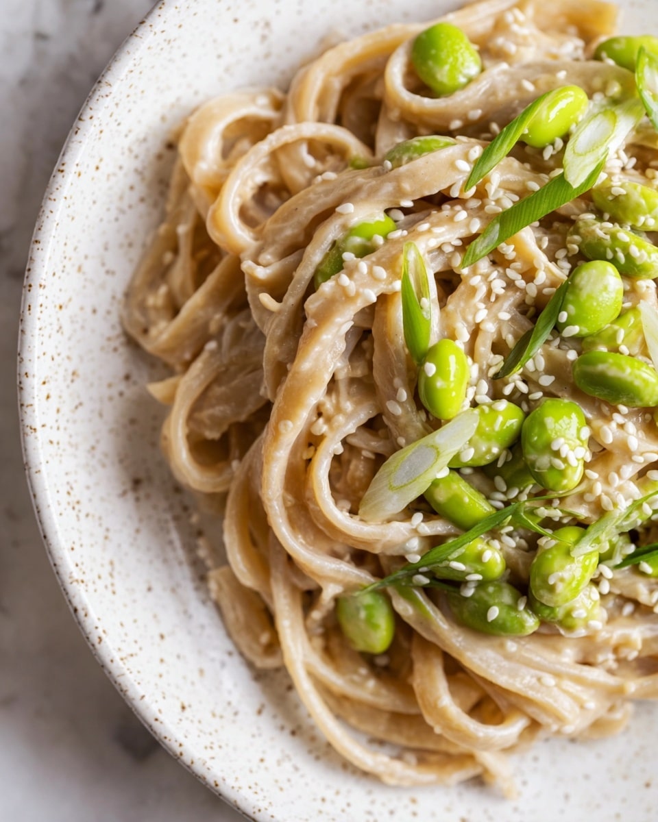 This dish shows a close-up of creamy, light brown noodles piled on the left side of a white speckled plate, with a smooth and slightly shiny texture. On the right, there is a topping of bright green edamame beans mixed with small thin light green onion rings scattered on top. White sesame seeds are sprinkled over both the noodles and the edamame, adding small white dots all over. The plate sits on a white marbled surface. Photo taken with an iphone --ar 4:5 --v 7