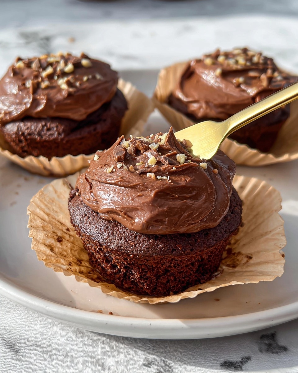 The image shows three chocolate cupcakes in light brown crinkled paper cups on a white plate with a white marbled texture surface beneath it. Each cupcake has a soft, dark brown base topped with a thick layer of smooth, shiny chocolate frosting sprinkled with small crumbs of nuts and chocolate bits. In the foreground, a golden knife is gently spreading the rich chocolate frosting over a cupcake. The background is softly focused, emphasizing the creamy texture and glossy shine of the frosting on the cupcakes. Photo taken with an iphone --ar 4:5 --v 7