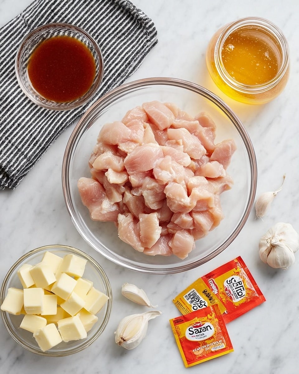 A clear glass bowl filled with light pink raw chicken pieces arranged in a loose mound at the center of a white marbled surface. To the upper left of the bowl is a small clear glass bowl with reddish brown sauce, placed on a black and white striped cloth. Below and left of the chicken bowl is another small clear glass bowl filled with pale yellow butter cubes. To the right of the butter are two bright orange seasoning packets labeled