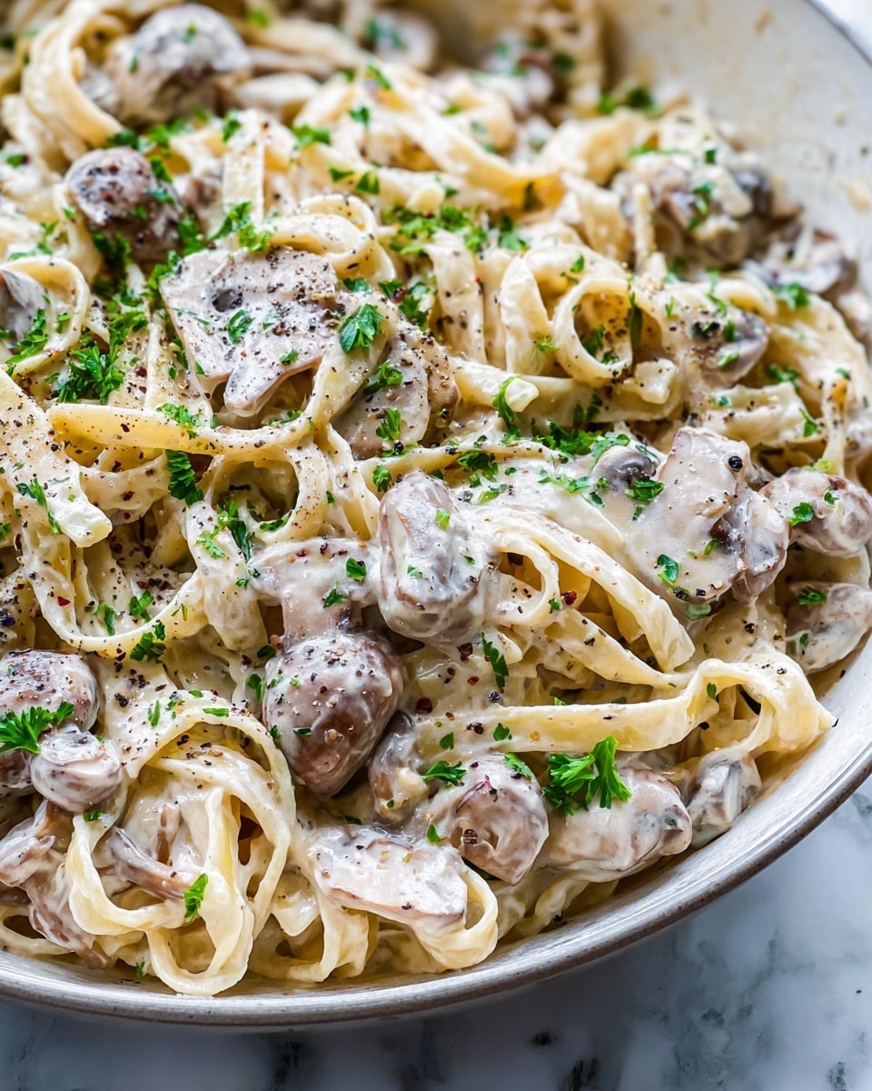 This image shows a close-up view of creamy mushroom pasta in a white bowl. The dish has multiple layers, starting with thick creamy white sauce covered noodles that look smooth and shiny. Scattered among the noodles are light brown, cooked mushroom pieces with a slightly soft texture. Fresh green parsley bits are sprinkled over the pasta, adding small pops of color. Tiny black pepper specs are visible throughout, giving the dish a seasoned look. The bowl rests on a white marbled surface, enhancing the fresh and clean presentation. photo taken with an iphone --ar 4:5 --v 7